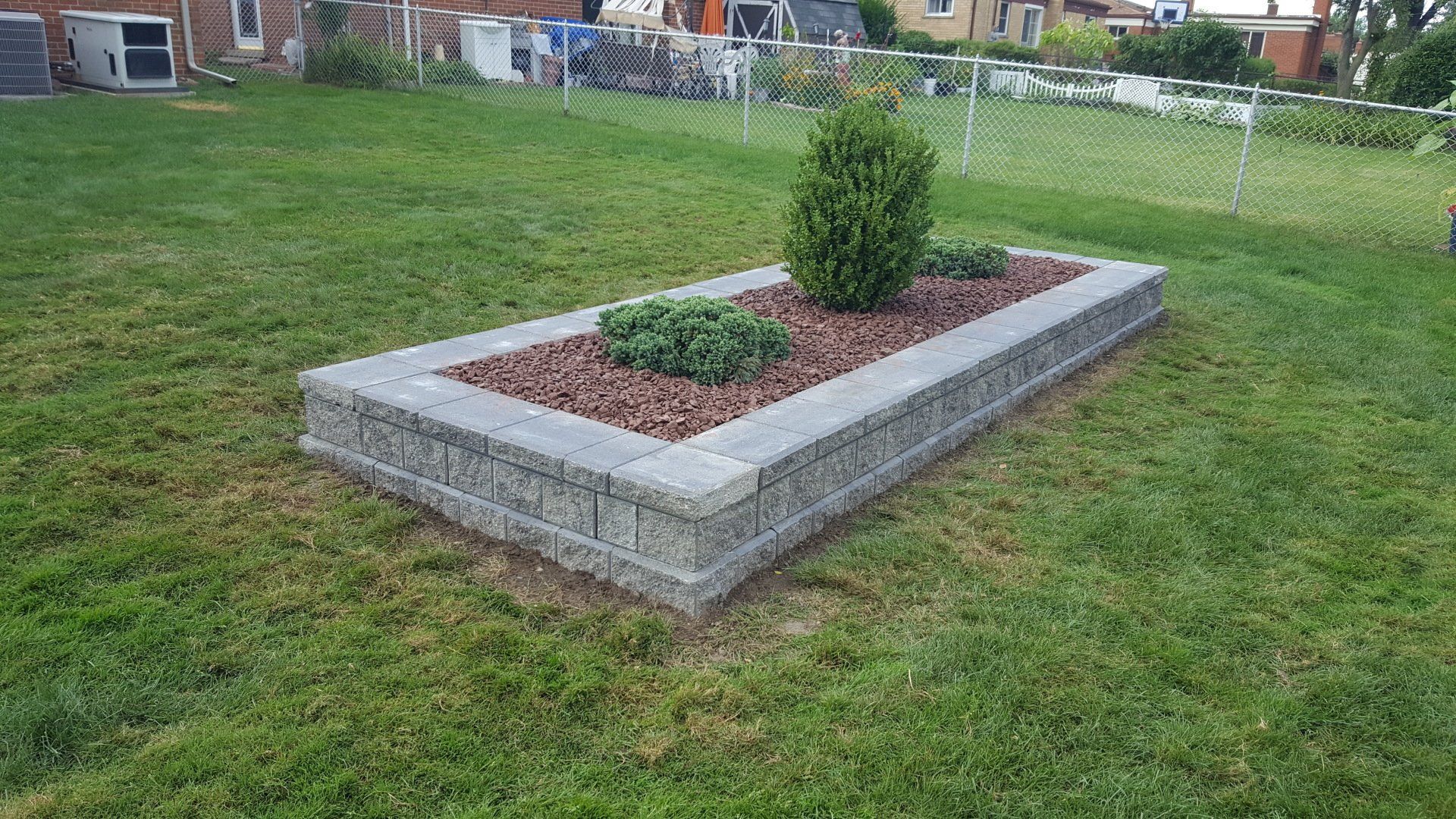 A rectangular raised garden bed made of grey stone blocks, containing two small green shrubs and reddish-brown mulch.