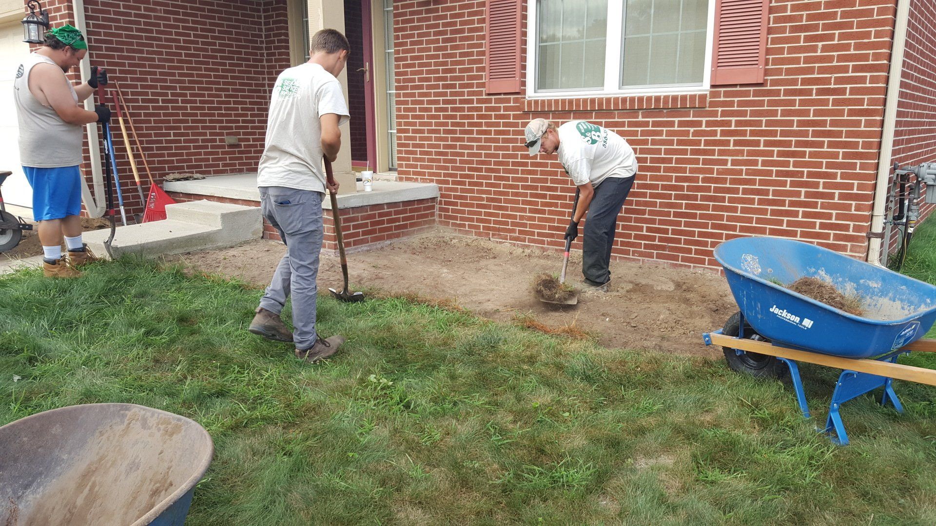 Three workers in casual clothes use tools to clear dirt and prepare the ground in front of a brick house.