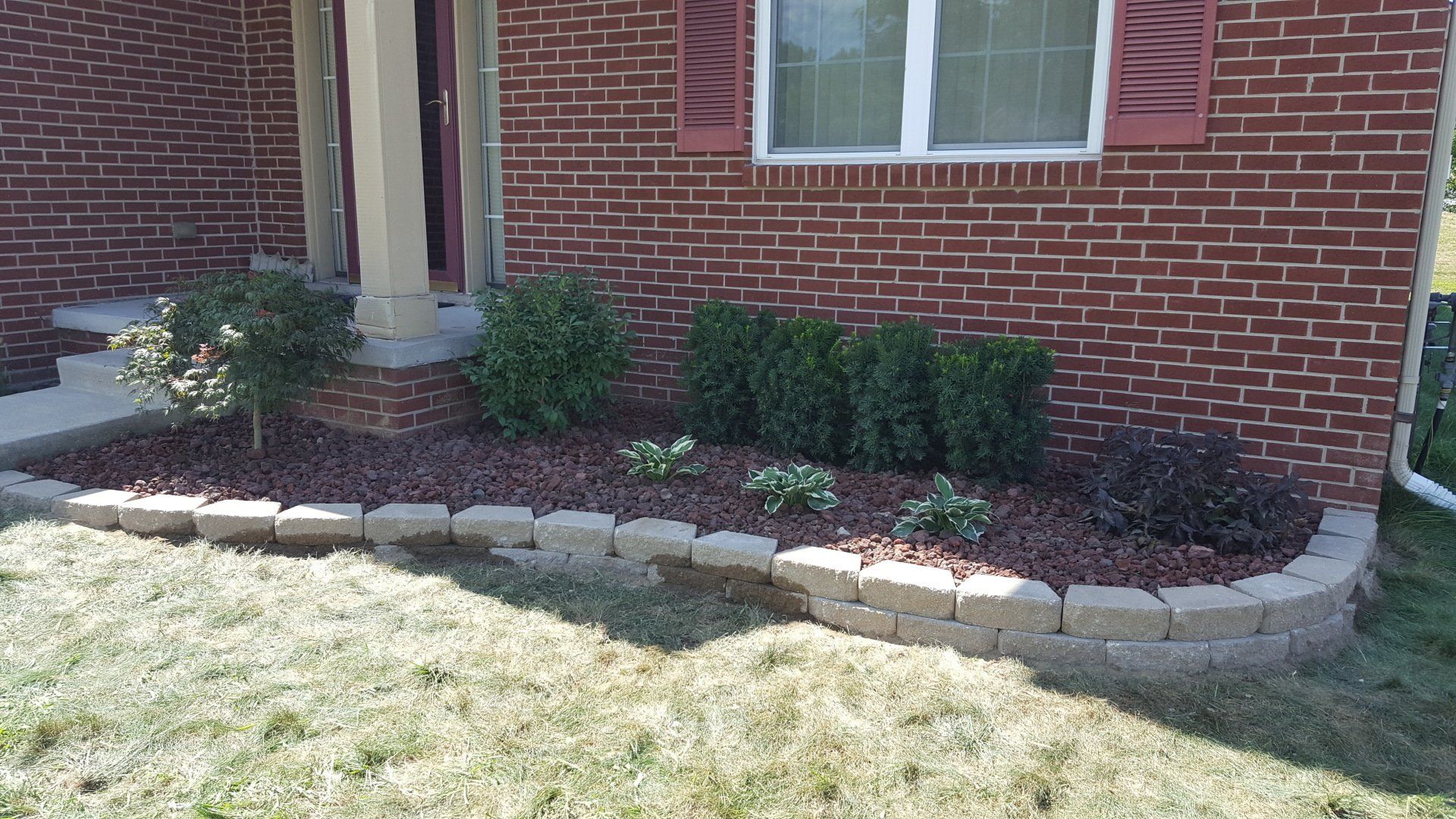 A brick house exterior with a curved, tiered stone retaining wall surrounding a mulch bed planted with various shrubs.