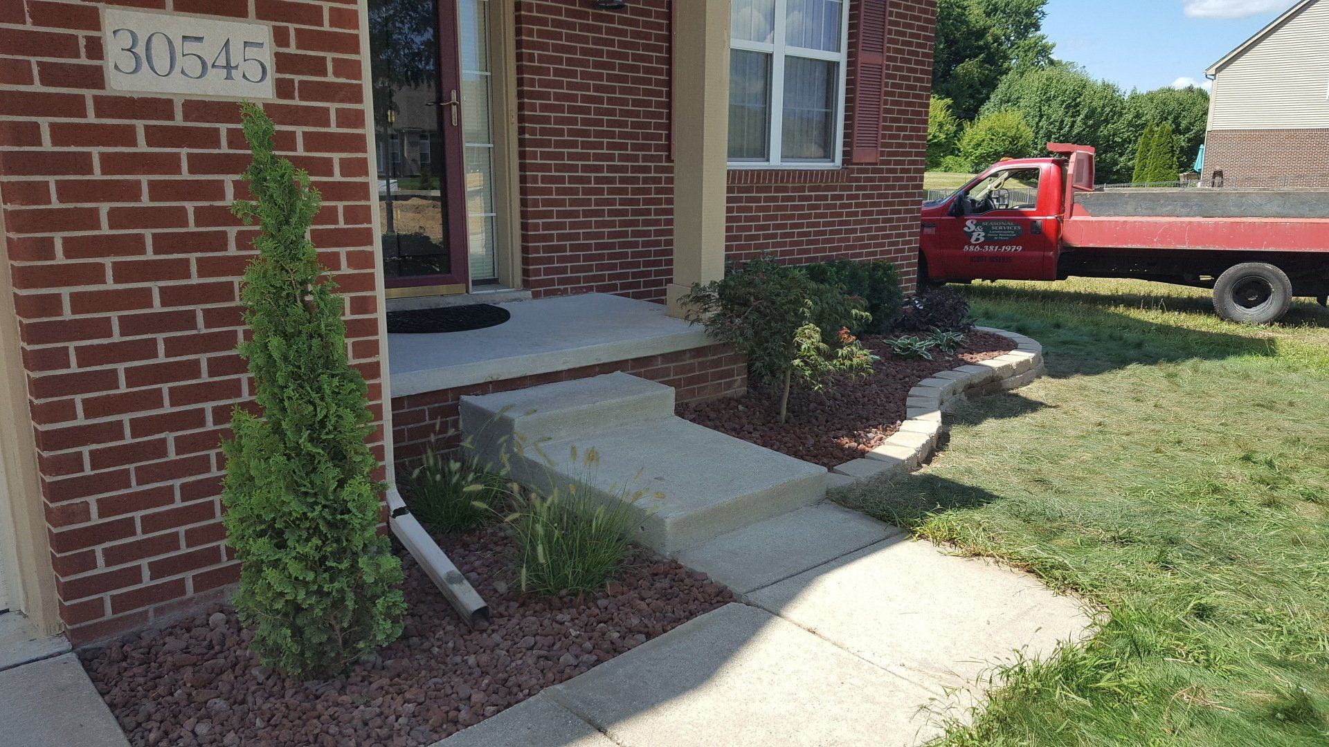 A brick house entrance with a concrete porch and steps, featuring a small evergreen tree and landscape mulch.