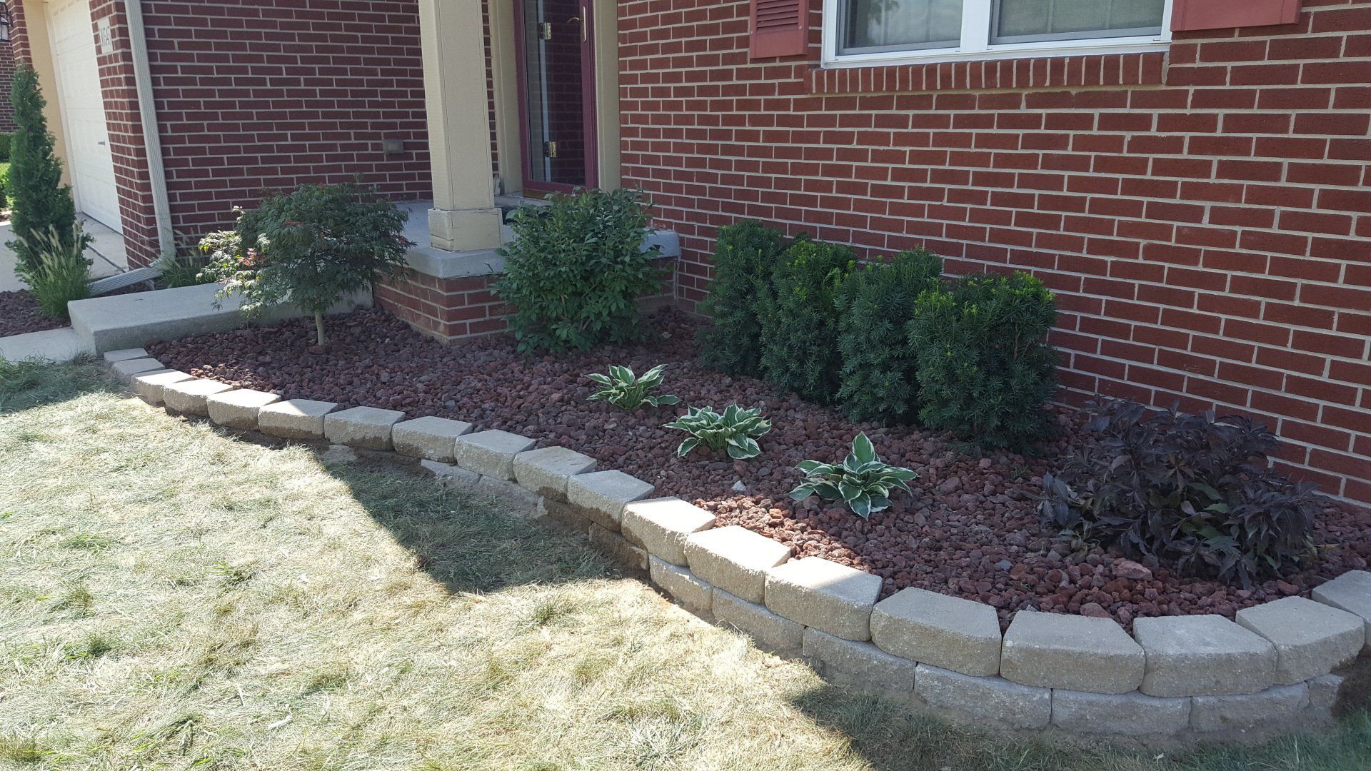 A curved, tan stone landscape border filled with reddish-brown mulch and small shrubs in front of a red brick house.