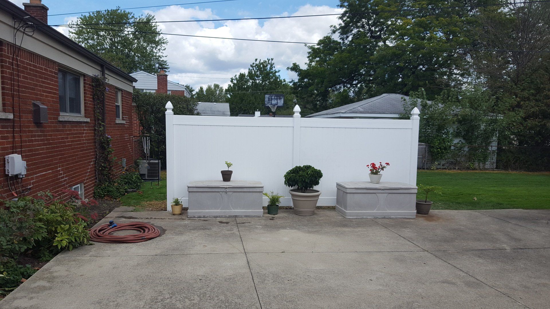 A white privacy fence stands in a backyard, featuring three storage chests with small potted plants on a concrete patio.