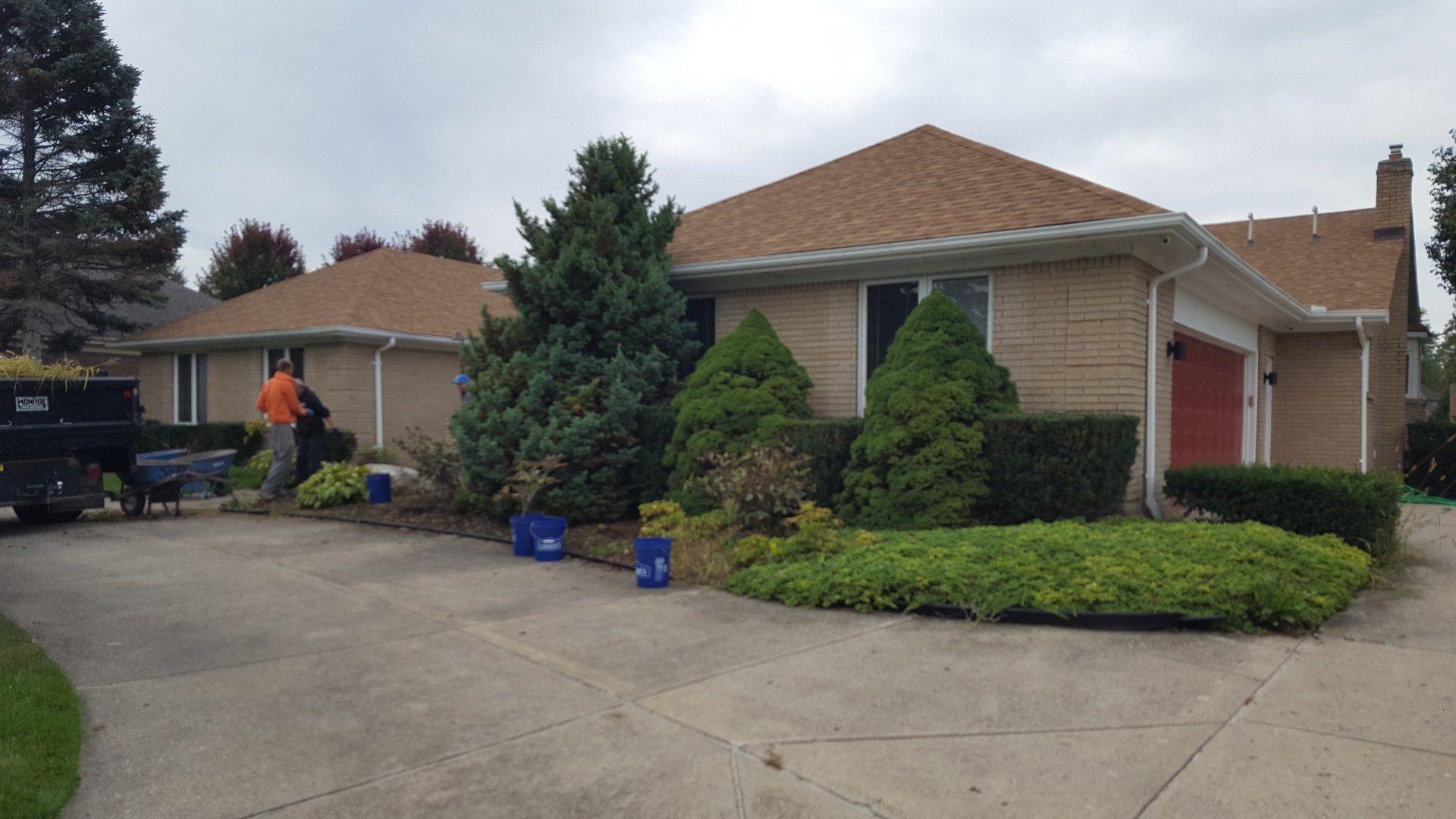 A person works in the landscaping of a tan brick house with a brown roof and a large concrete driveway.