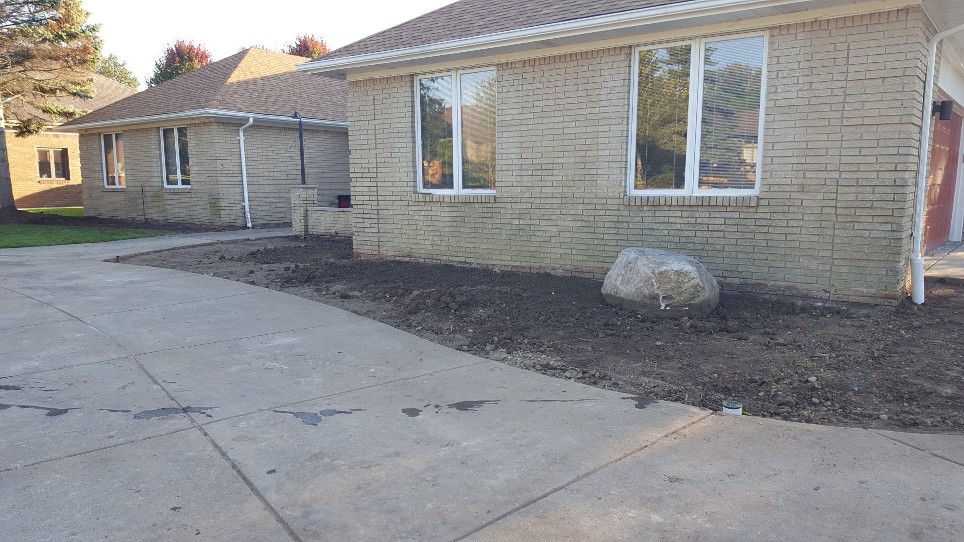 A beige brick house with a large boulder in the front landscaping bed next to a concrete driveway.