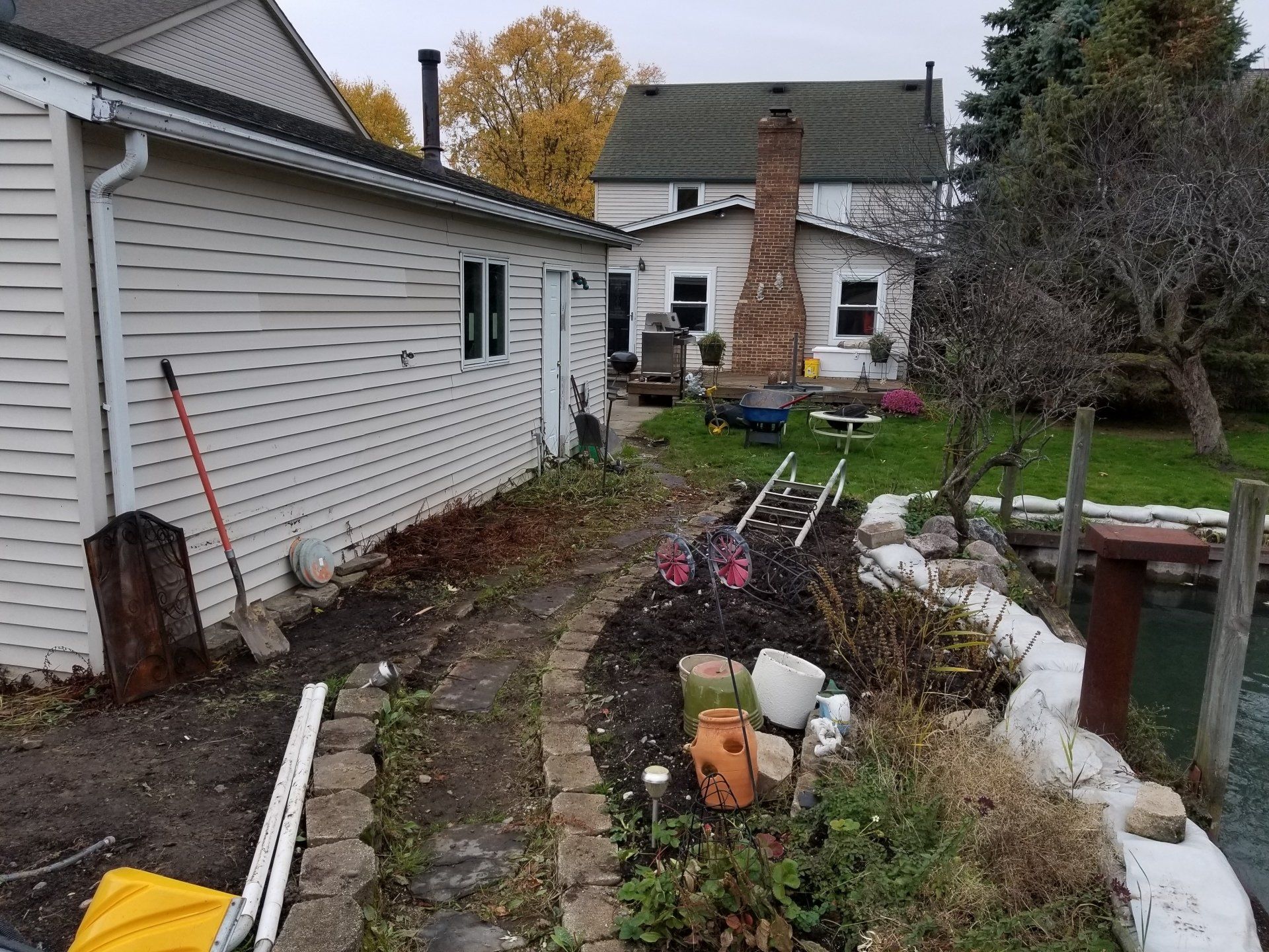 A side yard with a dirt path, a stone border, garden items, and houses in the background on an overcast day.