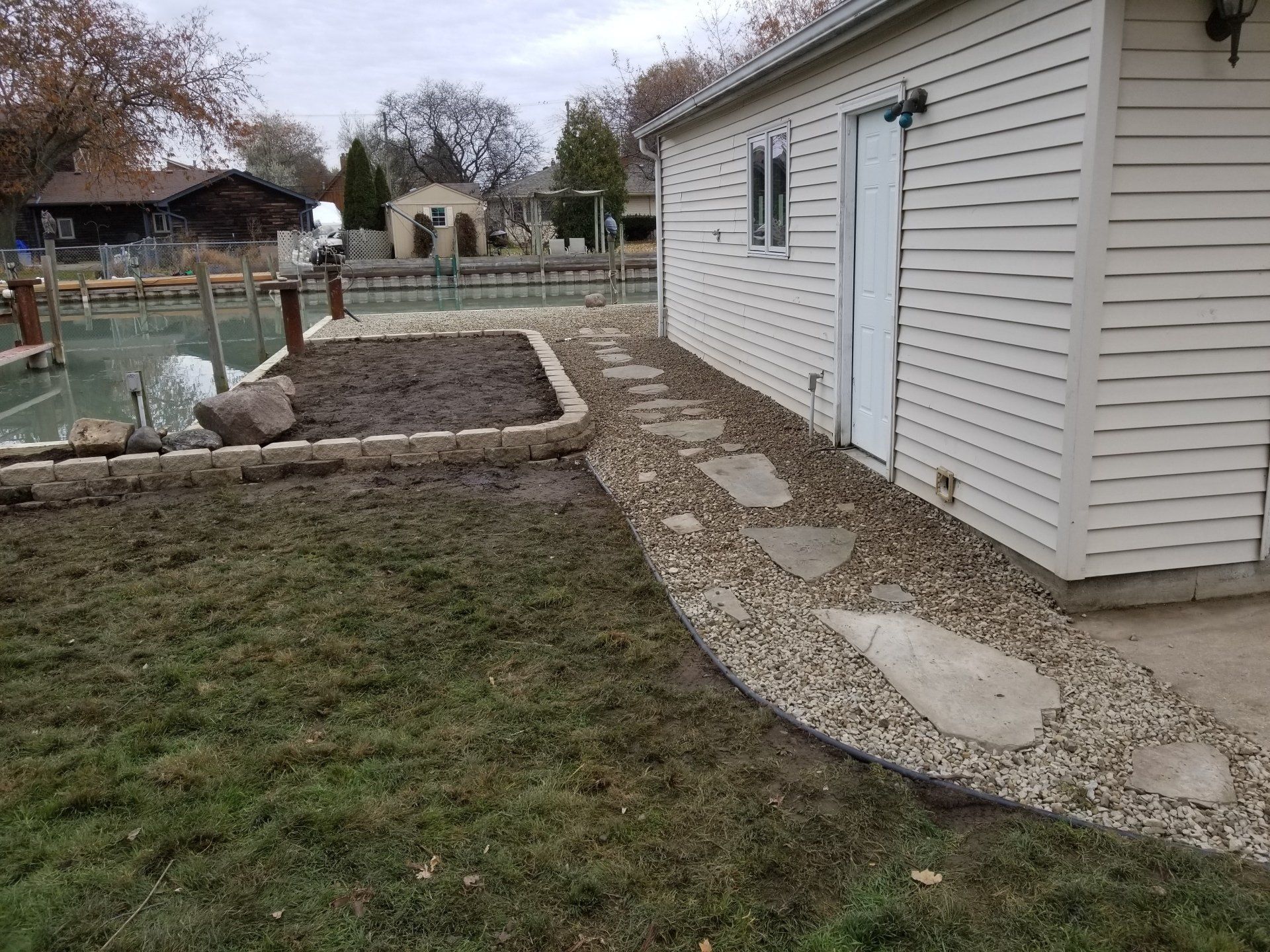 A side-yard view featuring a light-sided shed, a gravel path with stone pavers, and a dirt garden bed near the water.