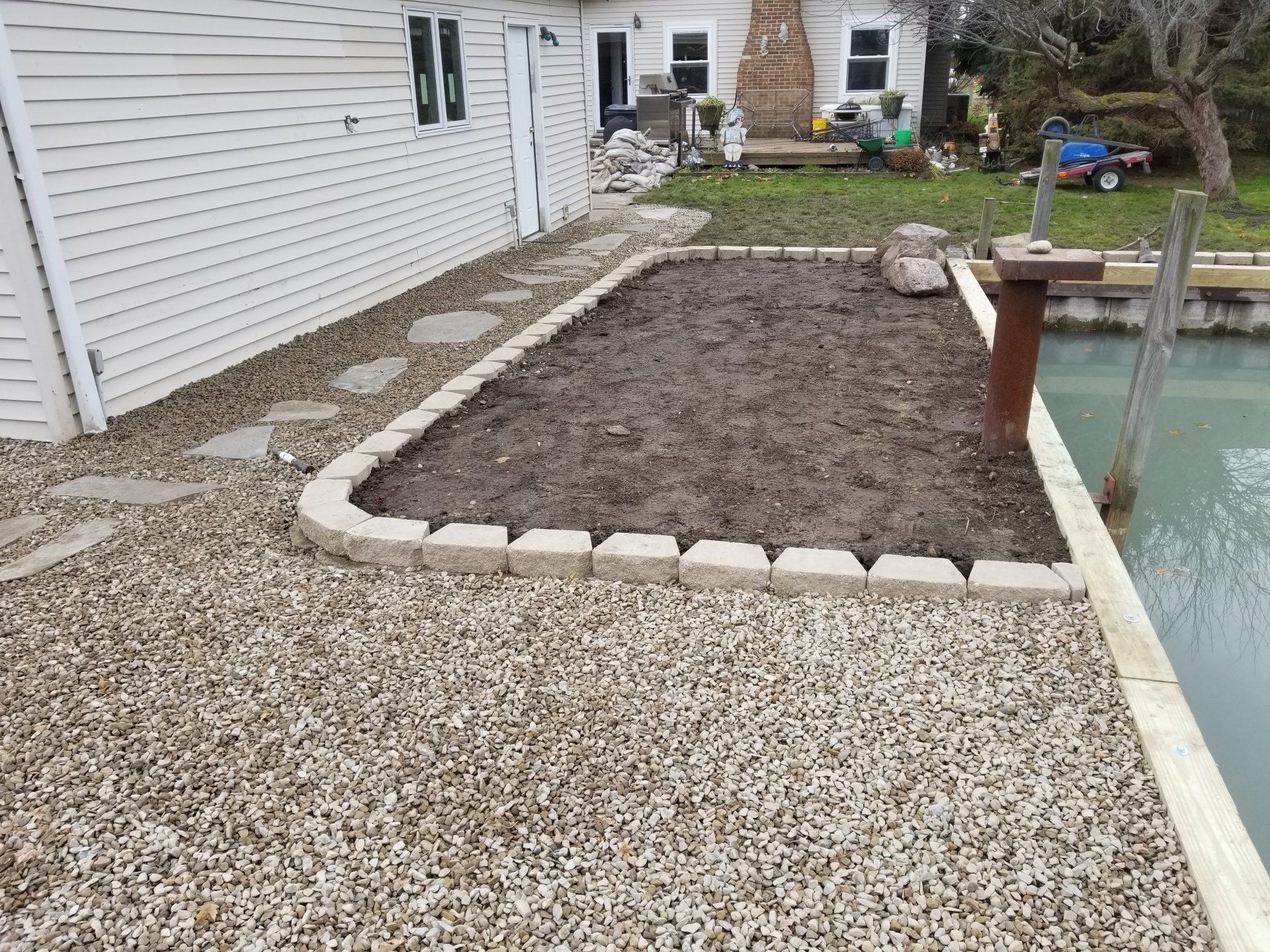 A rectangular garden bed with stone edging sits on a gravel shore next to a house and a small body of water.