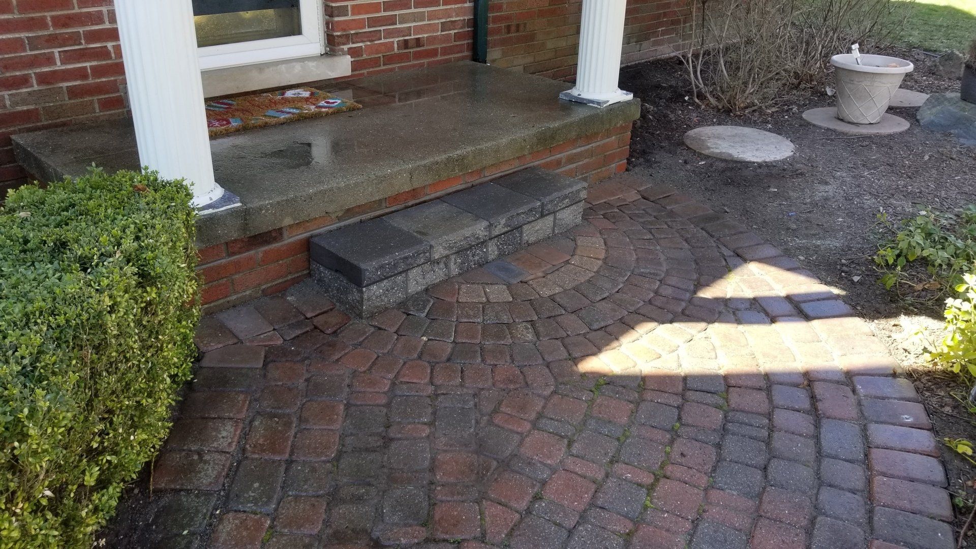 A brick house entryway with a stone porch step leading down to a semicircular patio made of interlocking red pavers.