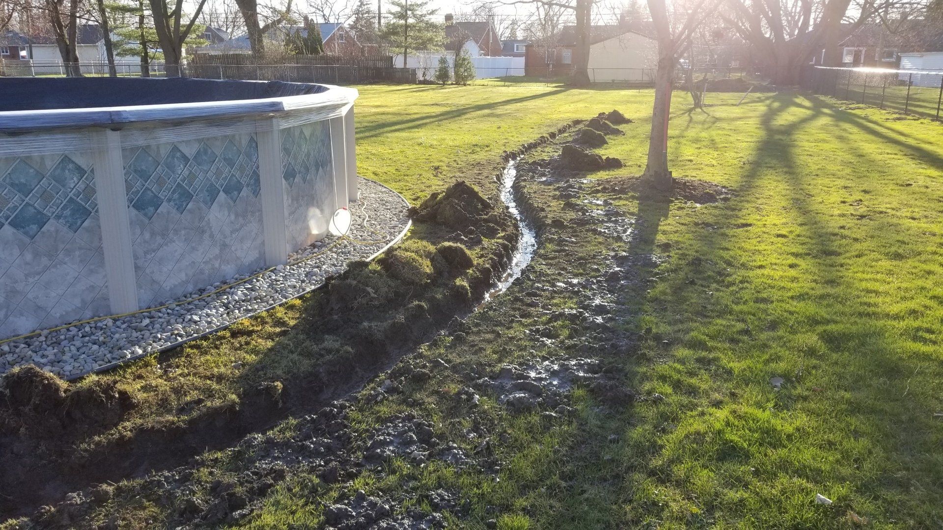 A trench dug alongside an above-ground pool in a grassy backyard during late afternoon.
