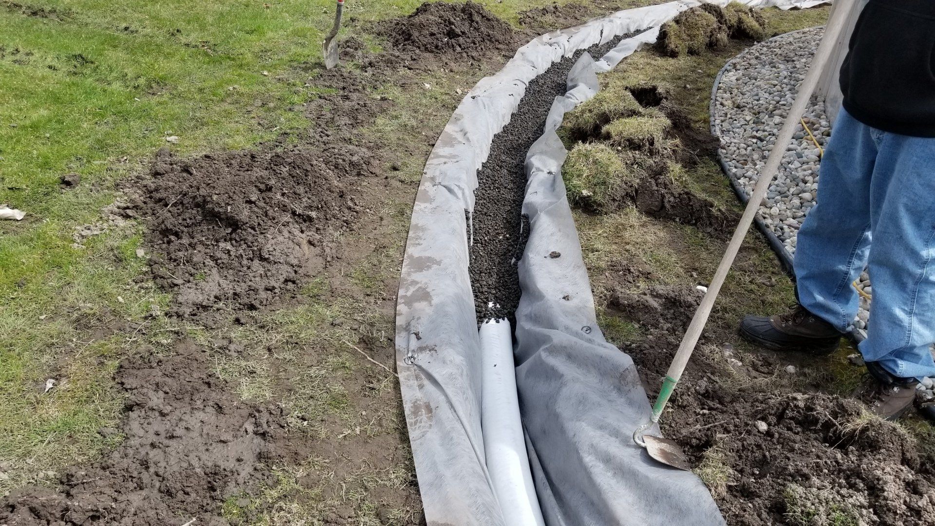A person uses a shovel to work on a French drain installation, placing gravel over a perforated pipe lined with fabric.