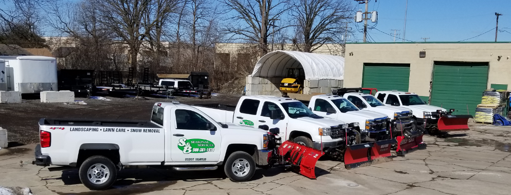 A row of white pickup trucks with snow plows attached, parked in a lot outside a commercial building.