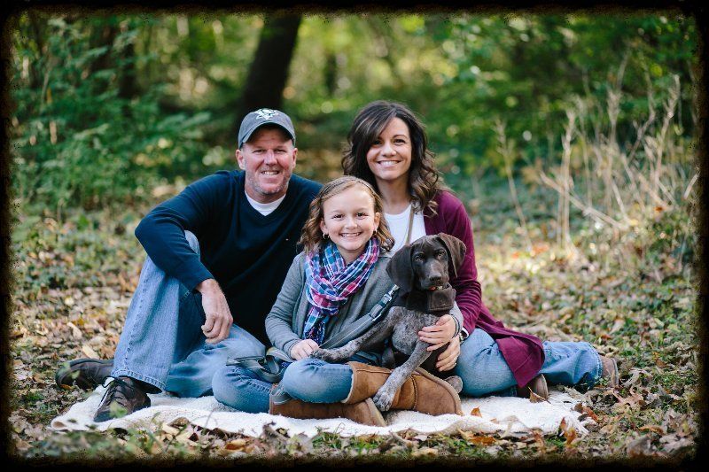 A family is sitting on a blanket in the woods with a dog.