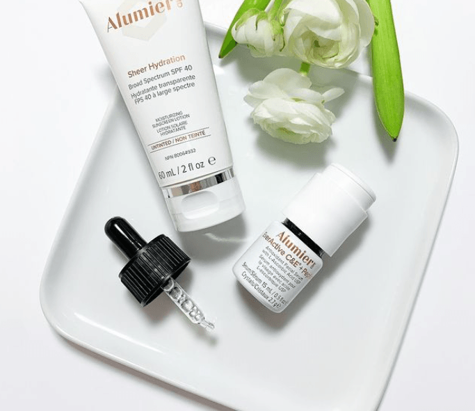 Skincare products on a white tray with white flowers.  One bottle has a dropper, the other is a tube. Alumier skincare brand.