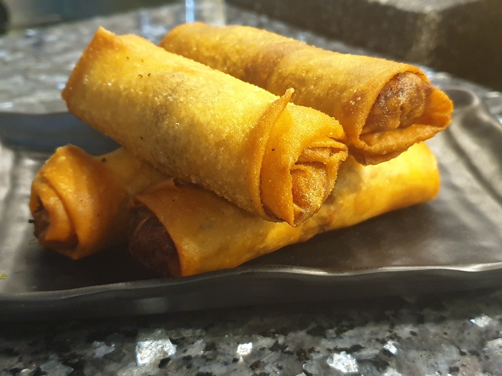 Three golden-brown fried egg rolls stacked on a square black plate set on a grey granite surface.