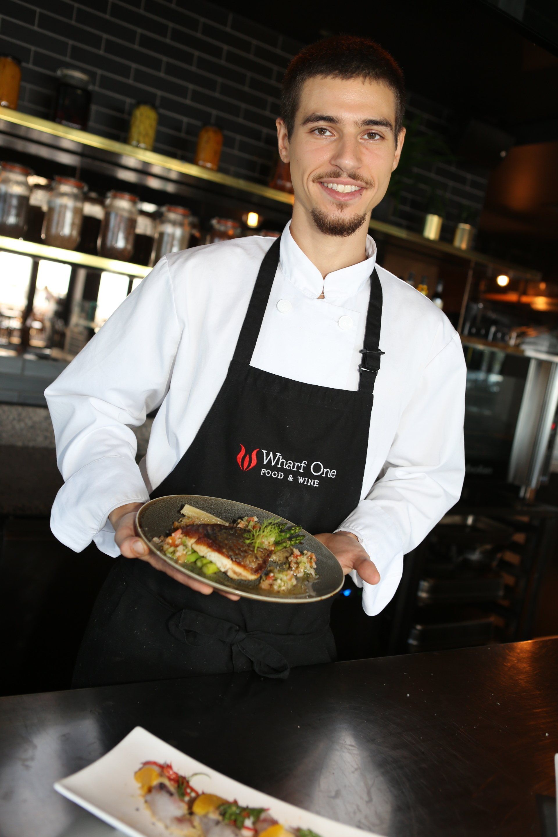 A chef in a white uniform and black apron holds a plate of food in a restaurant, with another dish on the counter below.
