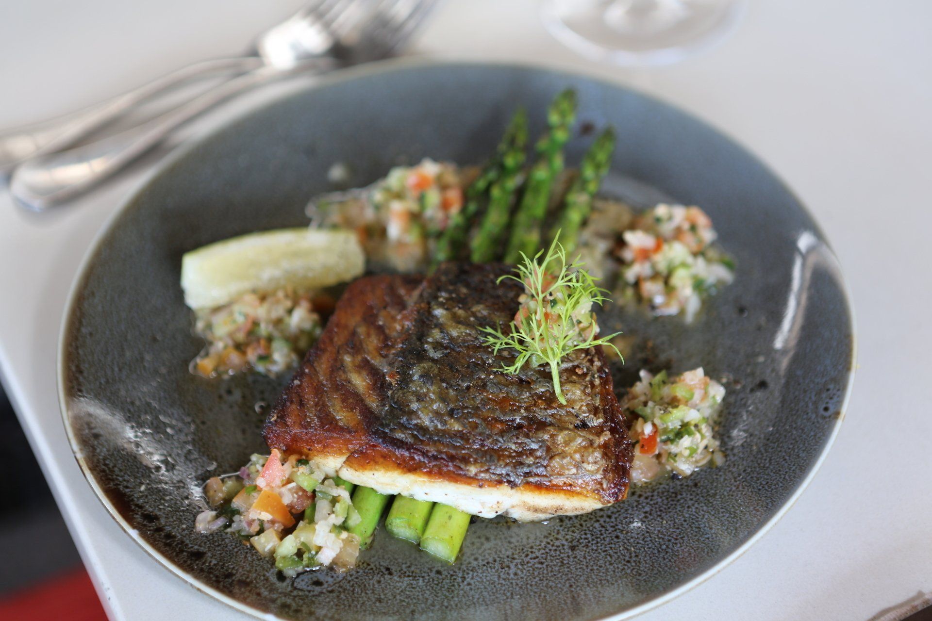 A seared fish fillet garnished with fresh herbs, served on a dark plate with asparagus and a chopped vegetable salsa.