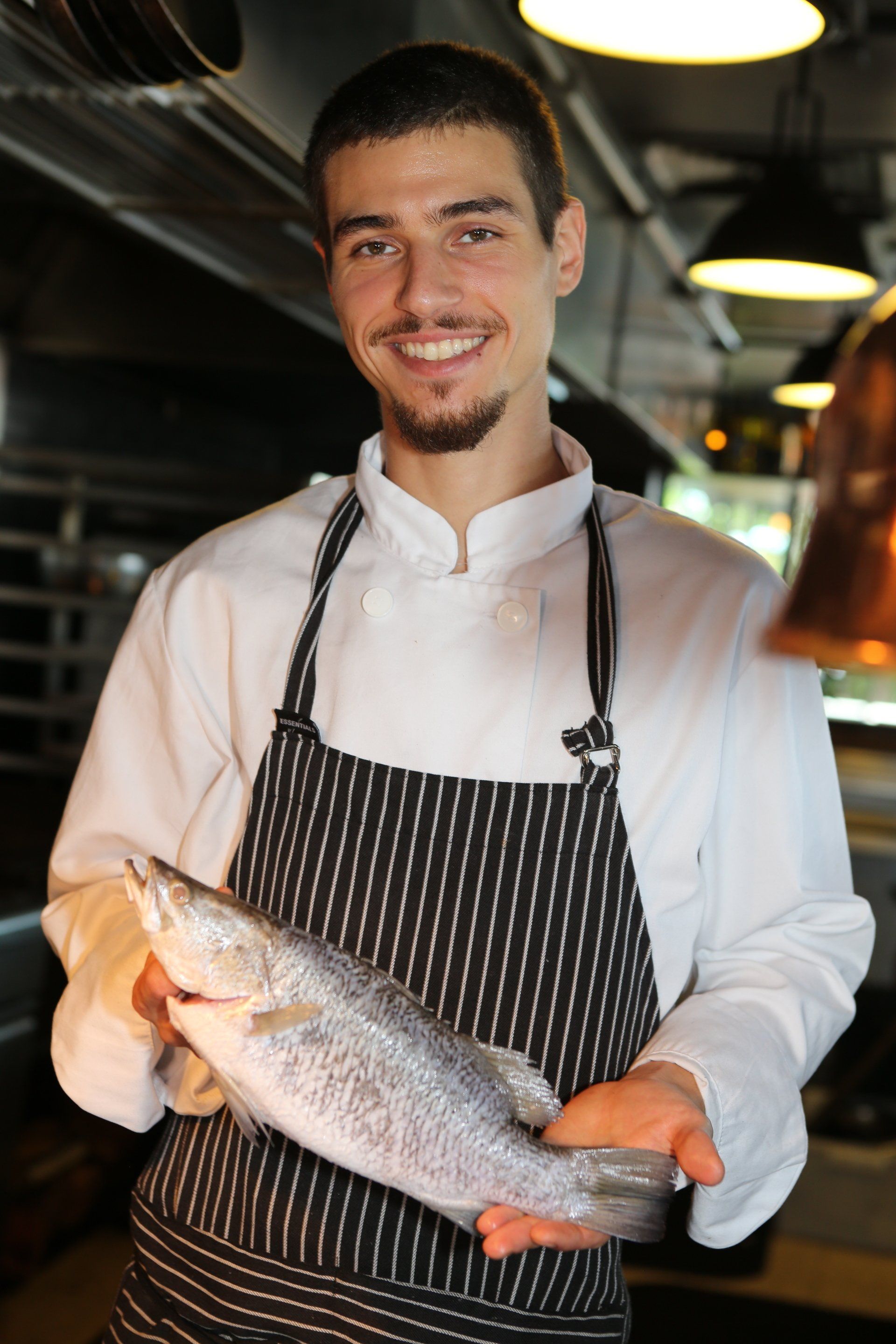 A smiling chef in a white uniform and striped apron holds a raw whole fish in a professional kitchen.