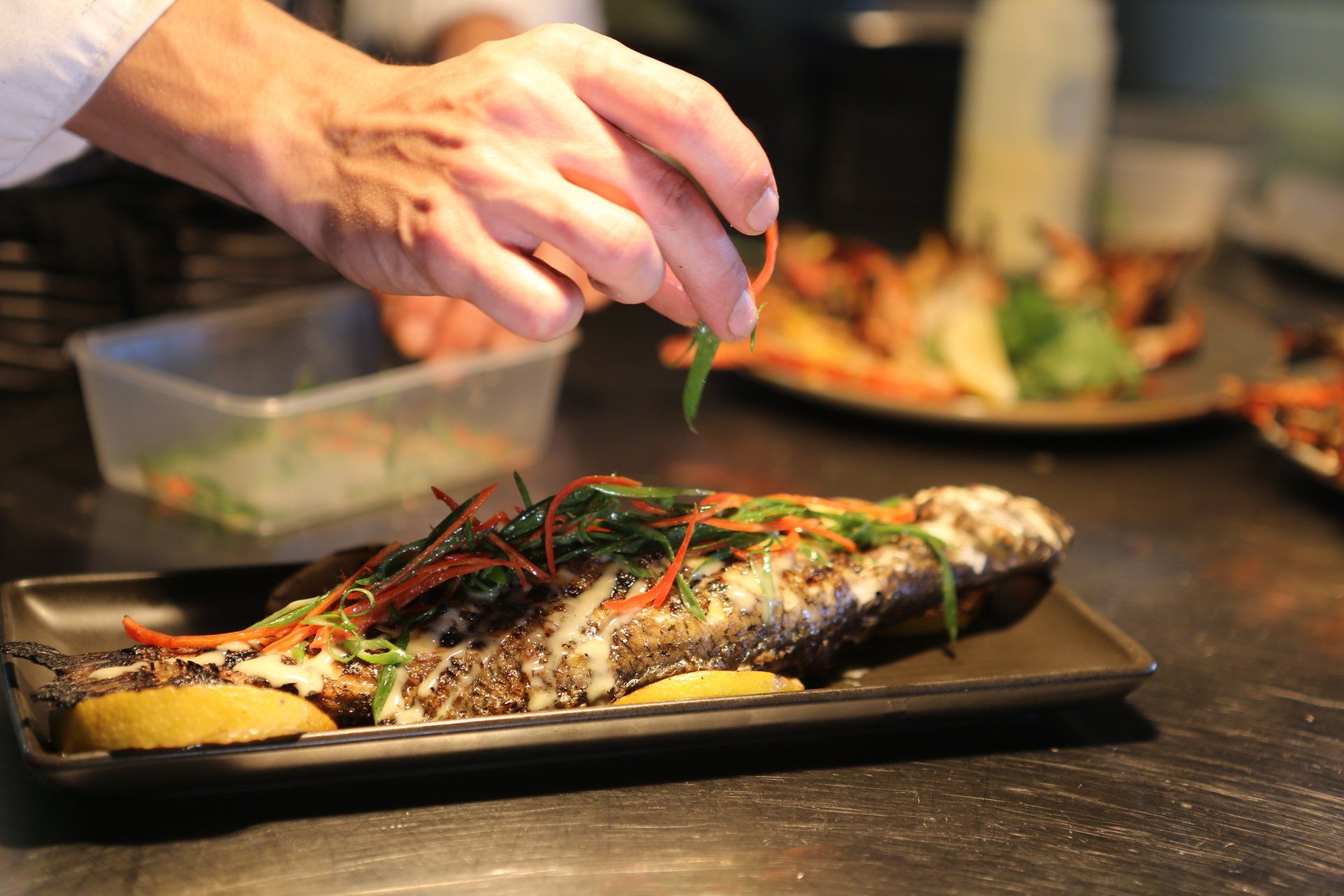 A chef garnishes a roasted whole fish on a rectangular platter in a professional kitchen.