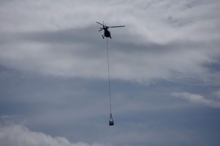A helicopter flying against a cloudy sky, carrying a suspended load via a long line.