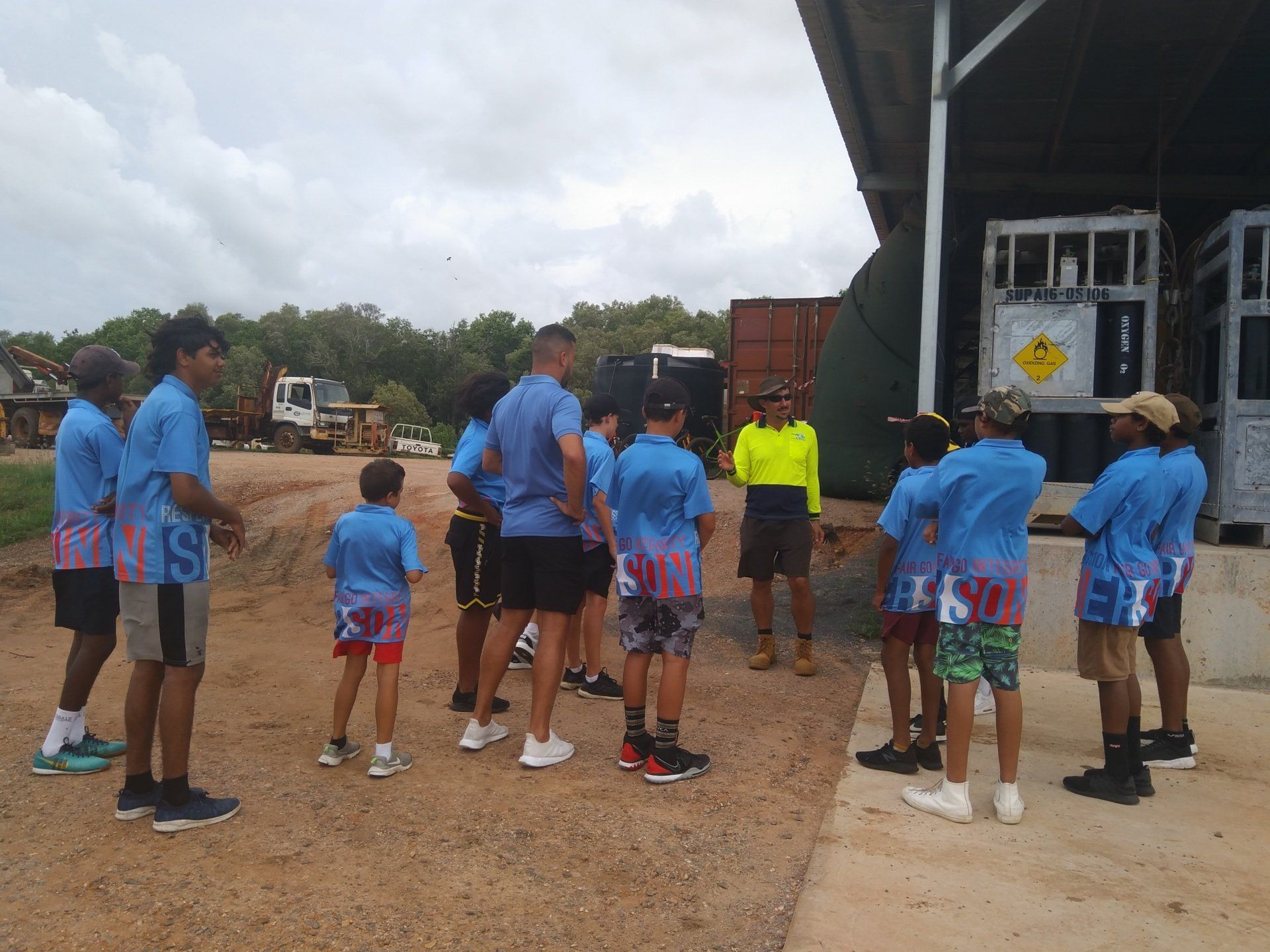 A group in matching blue uniforms stands on dirt, listening to a person in a high-visibility yellow shirt near a facility.