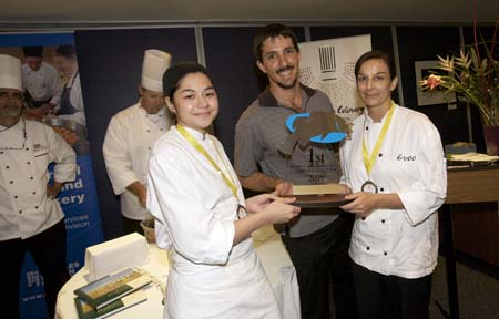 Two people in chef uniforms and a man holding an award trophy stand together at a culinary event.