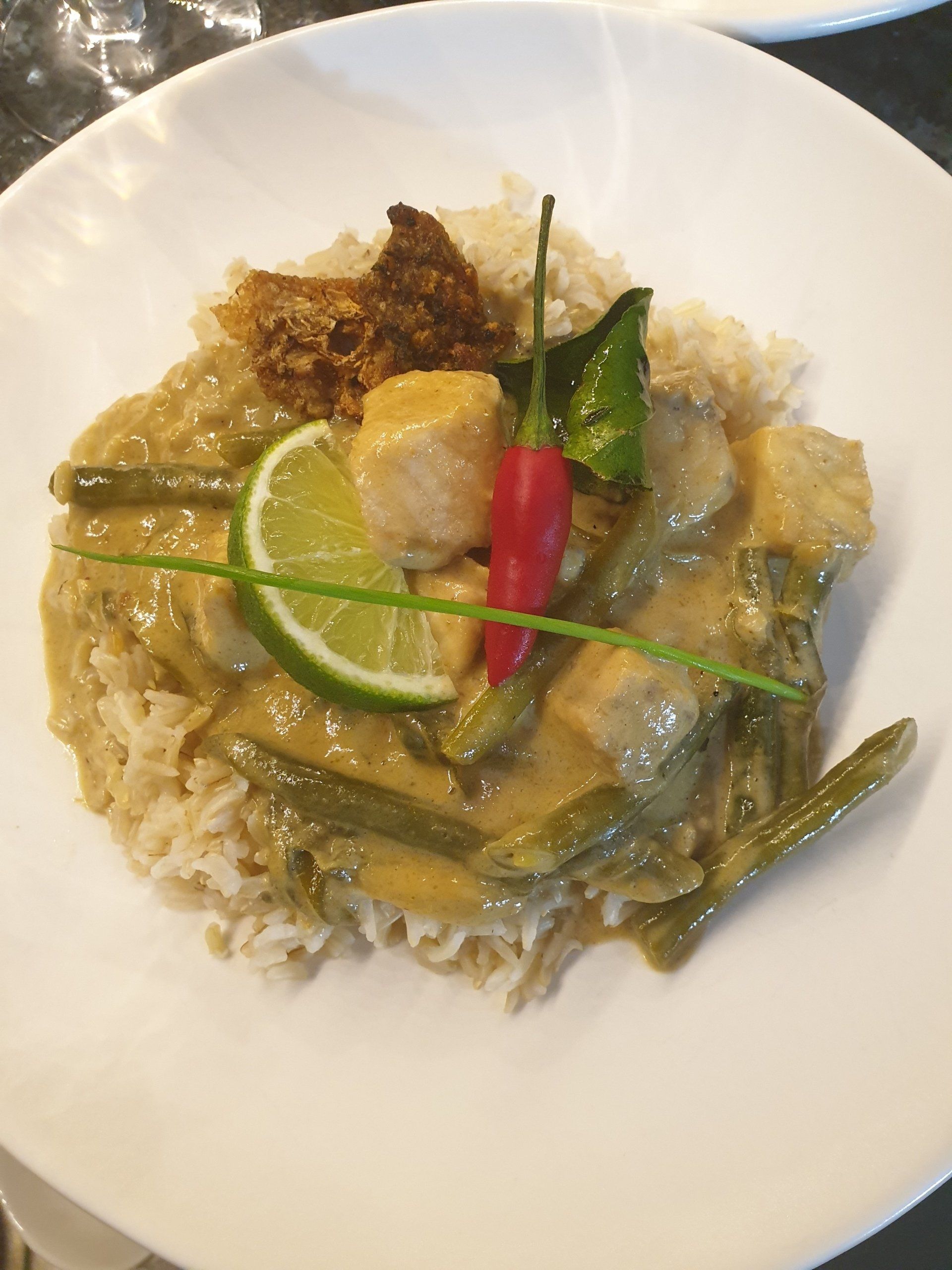 A plate of Thai green curry with chicken, green beans, and rice, garnished with a lime slice, a red chili, and herbs.