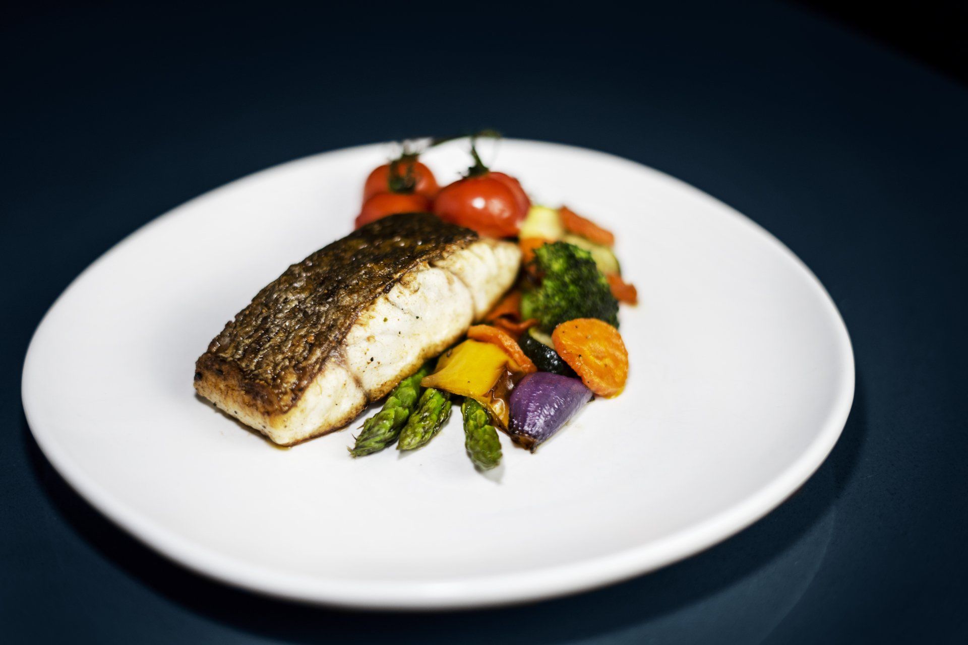 A seared fish fillet served on a white plate with roasted tomatoes, asparagus, broccoli, carrots, and onions.