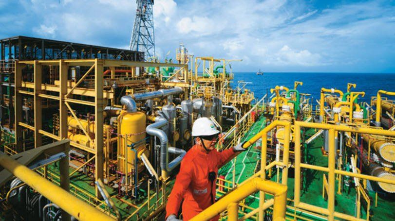 CONSULPRIME.COM - Trabalhador de macacão vermelho em uma plataforma de petróleo offshore, verificando equipamentos. Estruturas amarelas e cinzas, mar azul.