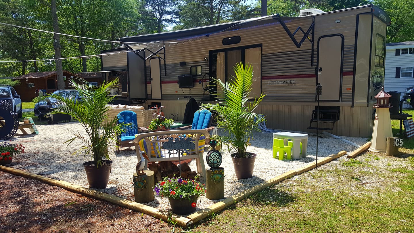 A rv parked in a gravel lot with a patio area surrounded by potted plants and chairs.