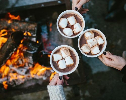 Three mugs of hot chocolate topped with marshmallows held near a campfire.