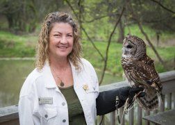 Woman in white jacket holds a barred owl outdoors near water.