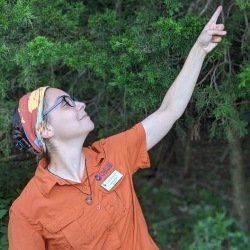 Woman in orange shirt points upward, looking up. She wears glasses, headband, and a nametag in a wooded area.