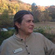 Woman with a name tag, wearing a button-down shirt, smiles outdoors with a fall foliage backdrop.