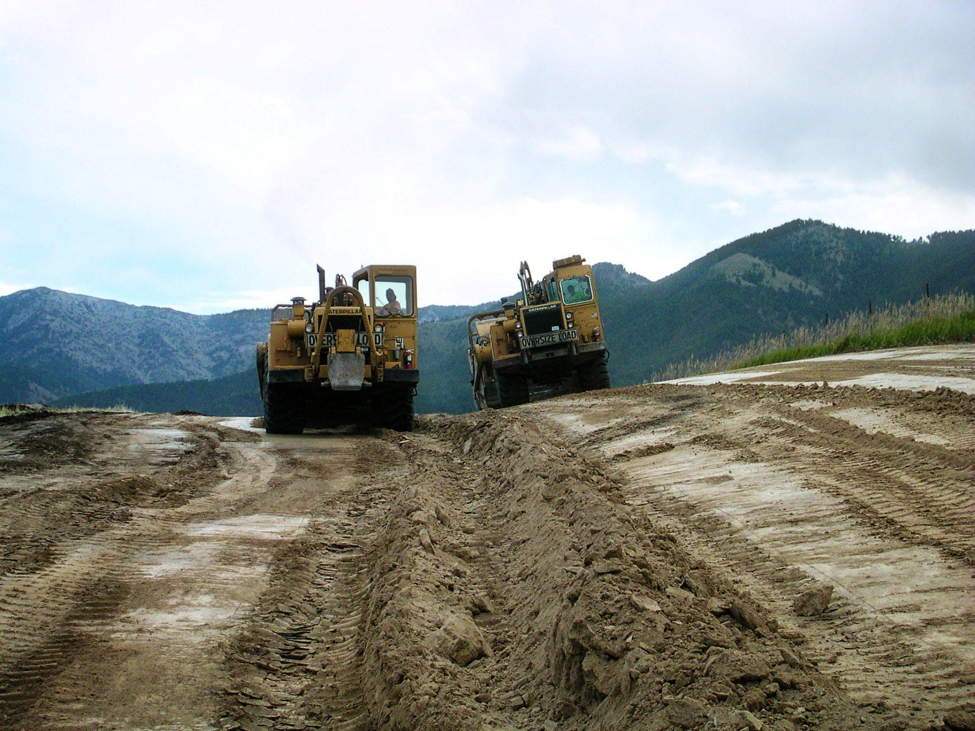 Two bulldozers are driving down a dirt road with mountains in the background