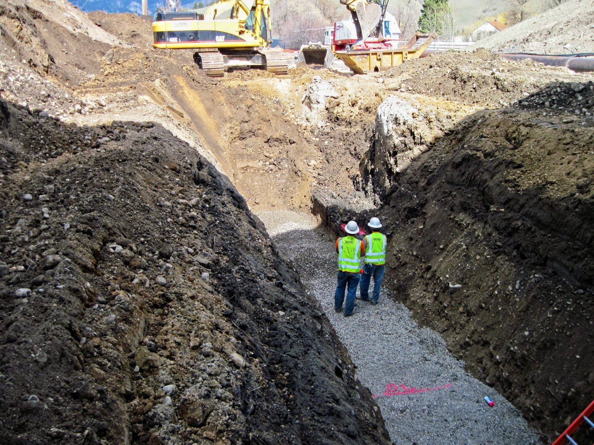 Two construction workers are standing in a trench looking at a pile of dirt.