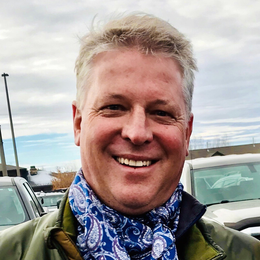 A man wearing a scarf and a jacket is smiling in a parking lot.