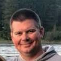 A man is smiling for a picture in front of a lake.