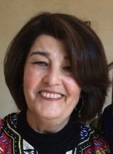 Woman with short, dark hair, smiling broadly. Wearing a patterned top and small earrings.