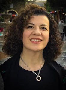 Woman with curly brown hair smiles, wearing a black top and silver necklace. Outdoors, blurry background.