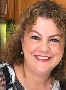 Woman with curly brown hair smiles, wearing a patterned top and pearl earrings, indoors.