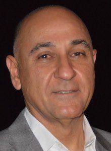 Bald man in a light suit, smiling. Lit from above. Dark background.