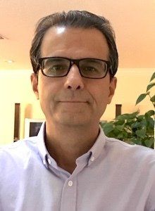 Man with glasses in a light blue shirt. Indoor setting with a plant visible behind him.