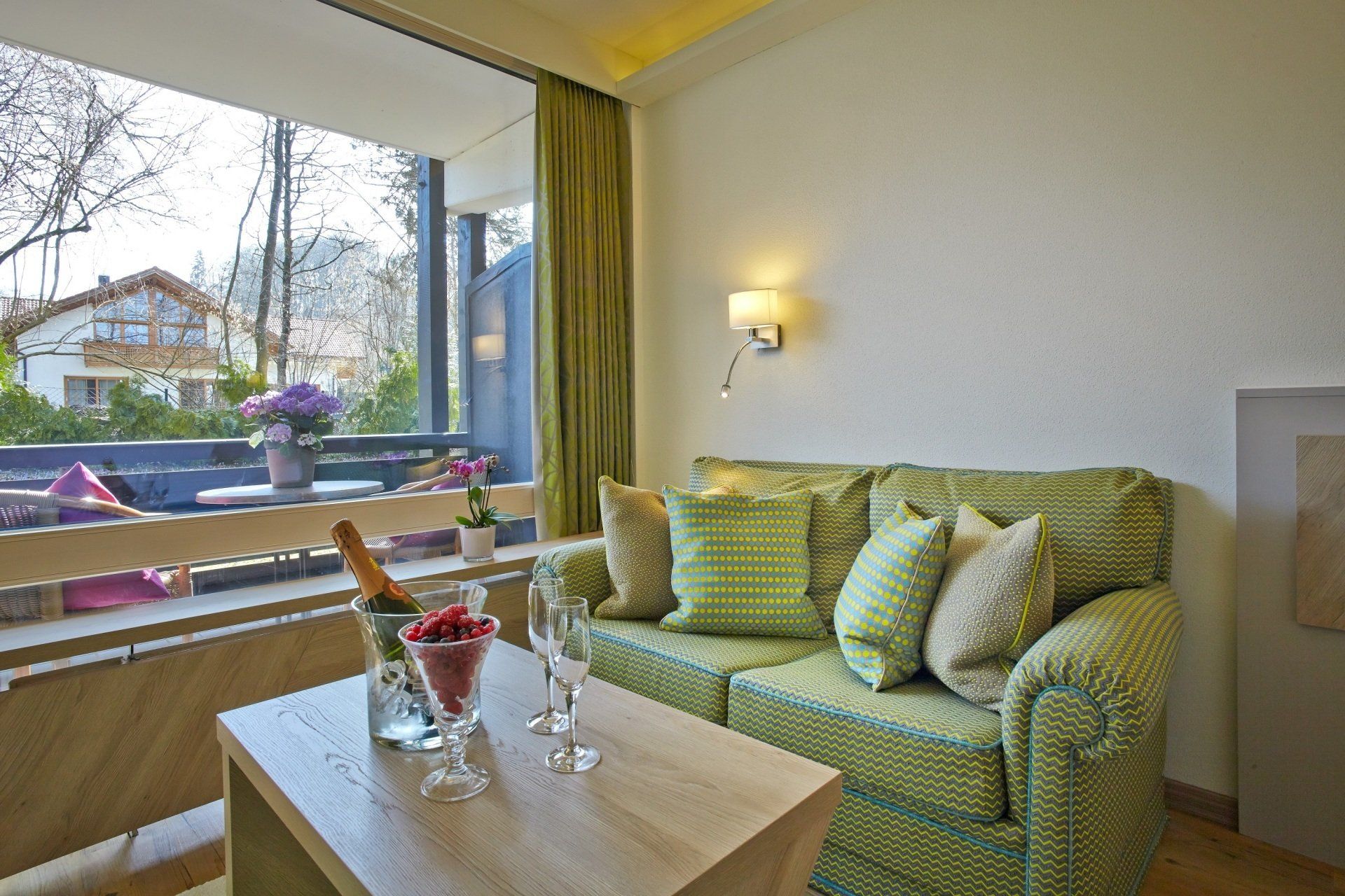Gemütliches Hotelzimmer mit grünem Sofa, Tisch, Sekt und Terrasse mit Blumen.