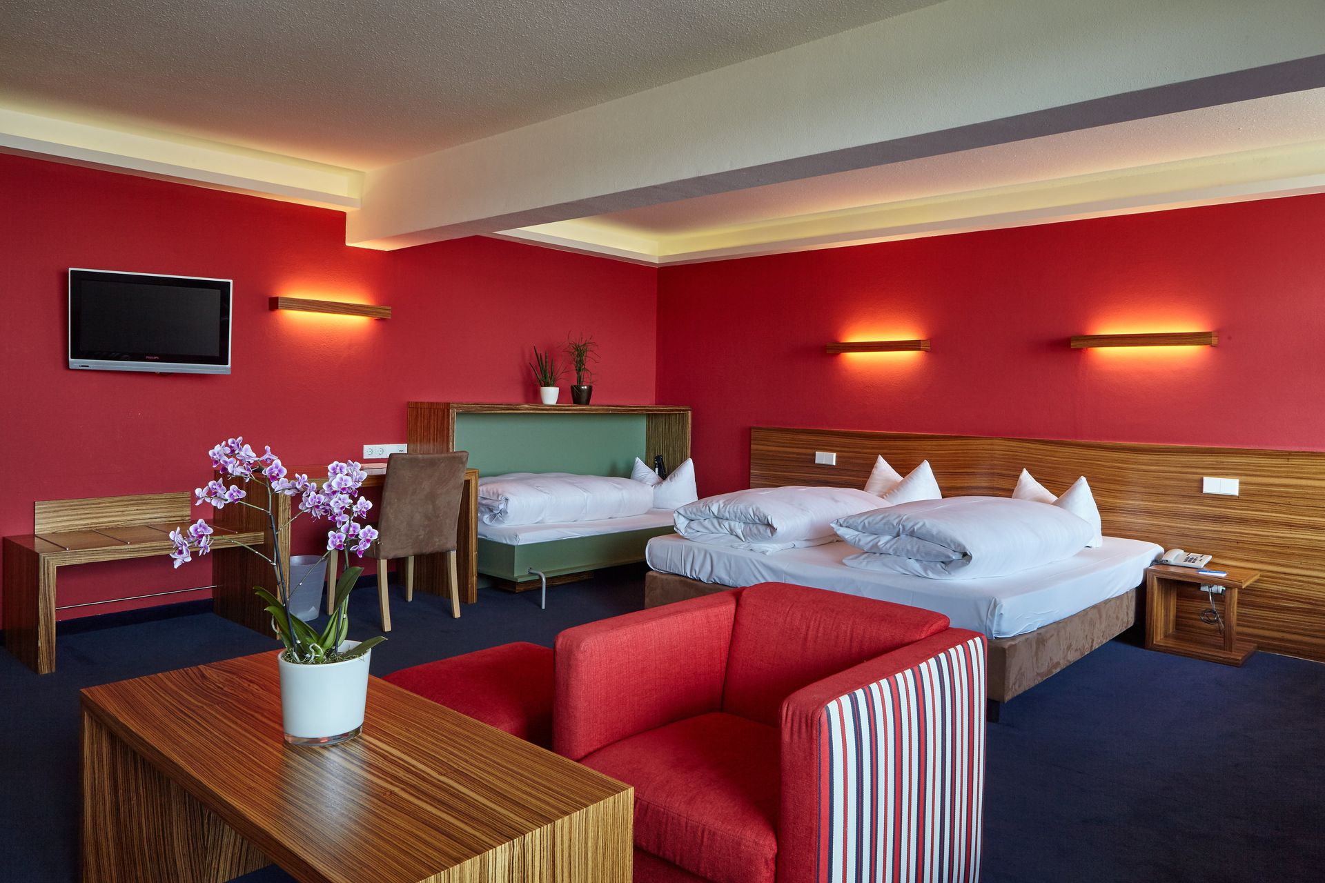 Gemütliches Doppelzimmer mit roter Wand, Sitzbereich, Orchidee auf Tisch und elegantem Bett.