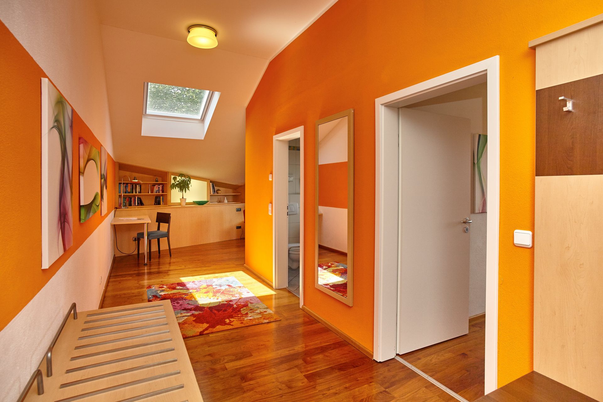 Heller Flur mit orangefarbener Wand, Spiegel, Schreibtisch unter Dachfenster und Bücherregal.
