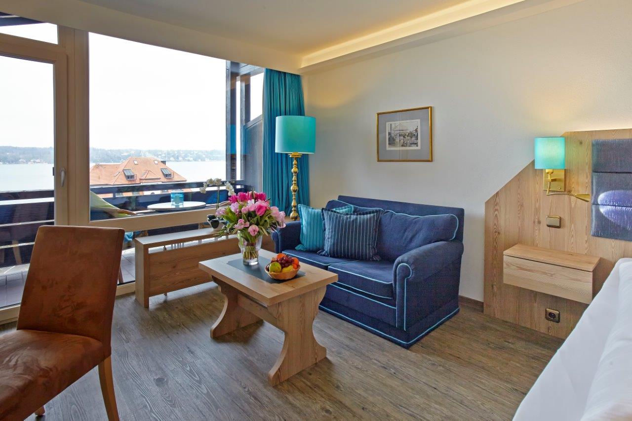 Hotelzimmer mit blauem Sofa, Holztisch, Blumenstrauß und Seeblick vom Balkon.