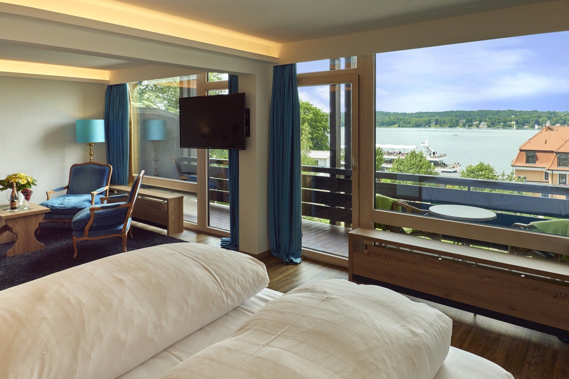 Hotelzimmer mit Doppelbett, blauen Sesseln, TV und Seeblick durch große Fensterfront.