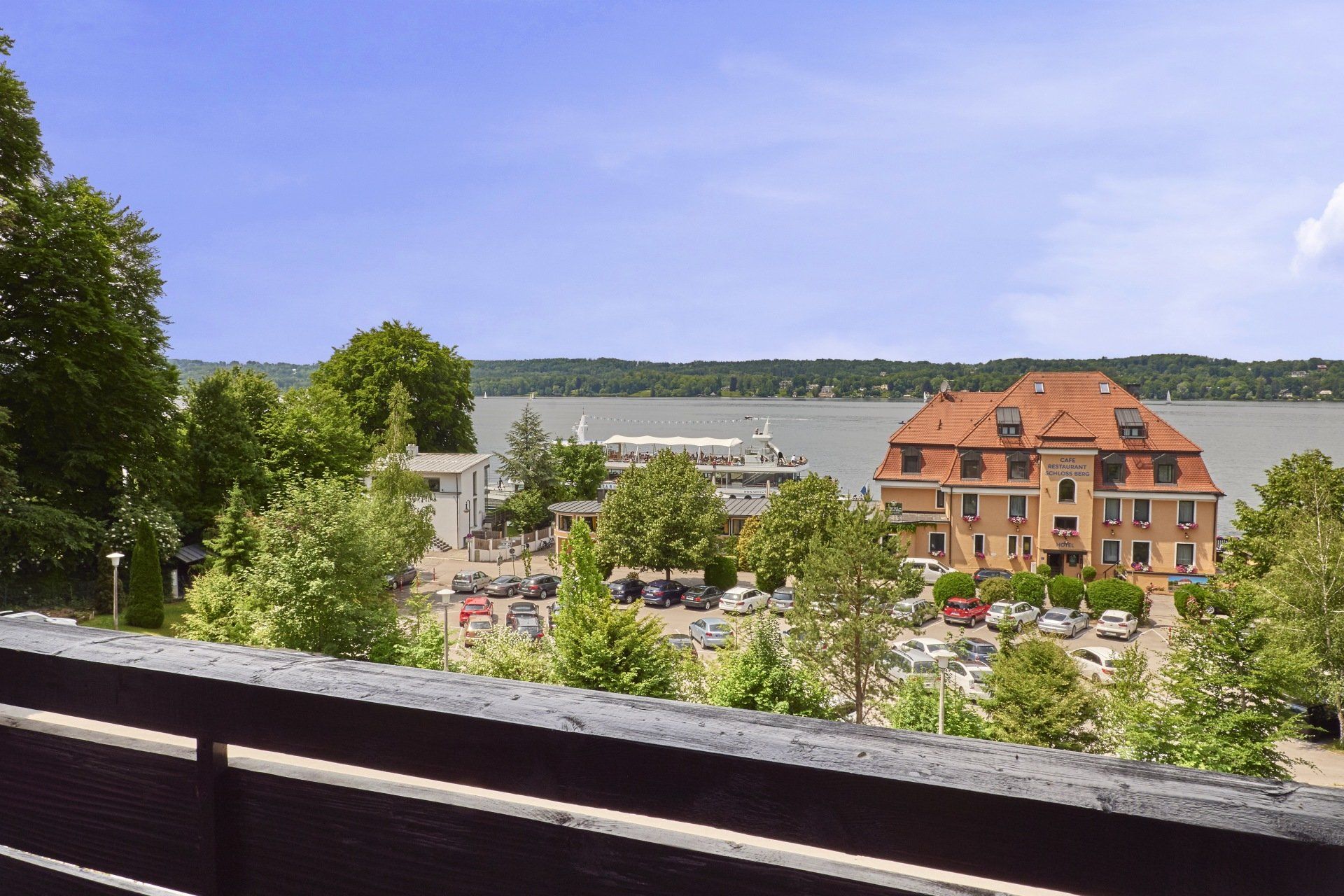 Blick vom Balkon auf See, Bäume, Hotelgebäude mit rotem Dach und Schiffsanleger.