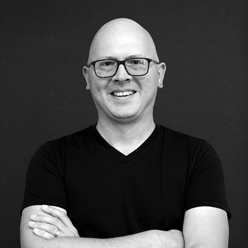 Bald man with glasses, arms crossed, smiling, wearing a black v-neck shirt against a dark background.