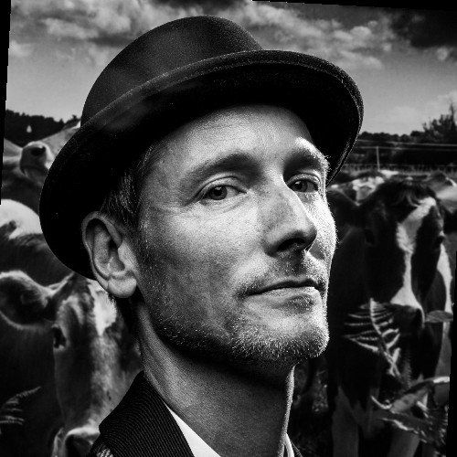 Man in hat, looking at camera, with cows in the background. Black and white.